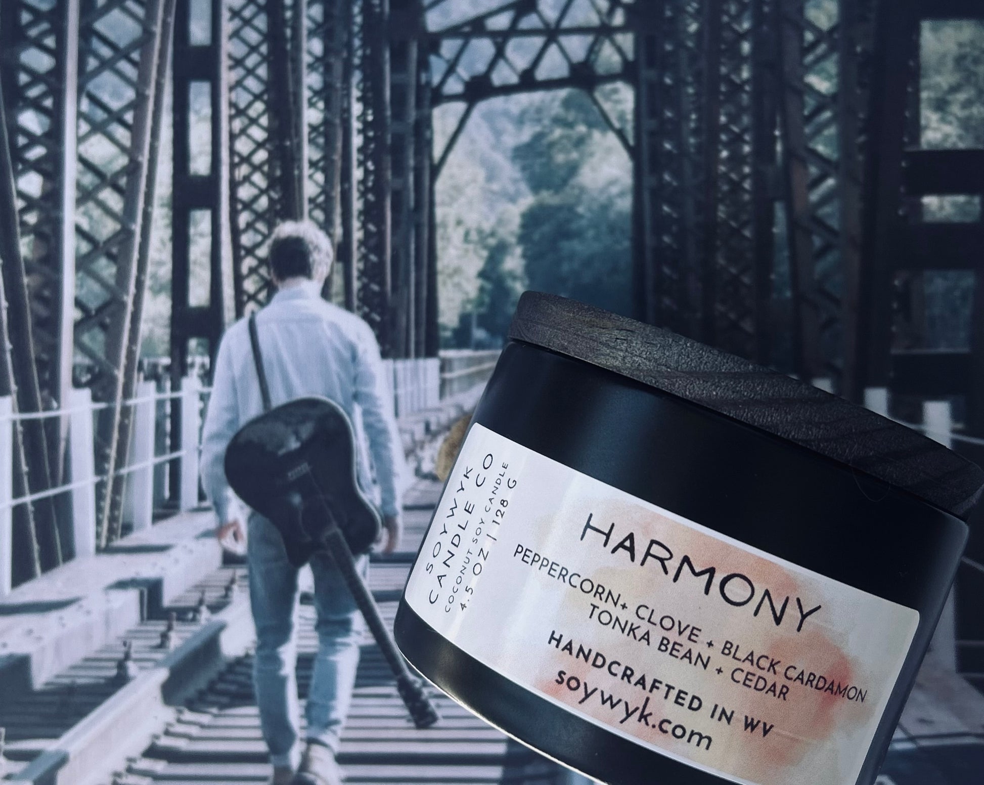 White Coconut Soy Candle in a modern 5 oz black  reusable ceramic jar  with a wood lid labeled "Harmony" with a young guy walking away from you across  a bridge with guitar strapped across his back. Featured fragrance note snare peppercorn, clove, black cardamon, tonka bean, cedar. One of the Signature Collections from Soywyk Candle Co. Hand-pour in WV. 