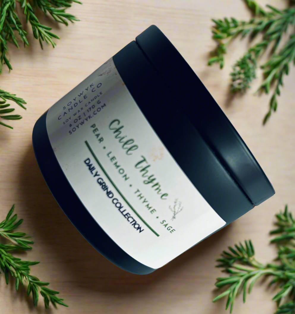 Soy wax candle in a 6 oz modern black candle tin labeled “ Chill Thyme “ on a beige countertop surrounded by herbs. Featured fragrant notes are pear, lemon, thyme, sage. One of the Daily Grind Collection by SoyWyk Candle Co, Hand- poured in WV. 