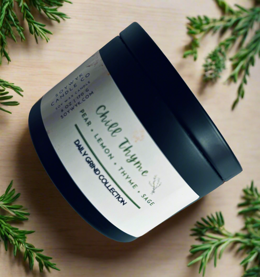 Soy wax candle in a 6 oz modern black candle tin labeled “ Chill Thyme “ on a beige countertop surrounded by herbs. Featured fragrant notes are pear, lemon, thyme, sage. One of the Daily Grind Collection by SoyWyk Candle Co, Hand- poured in WV. 