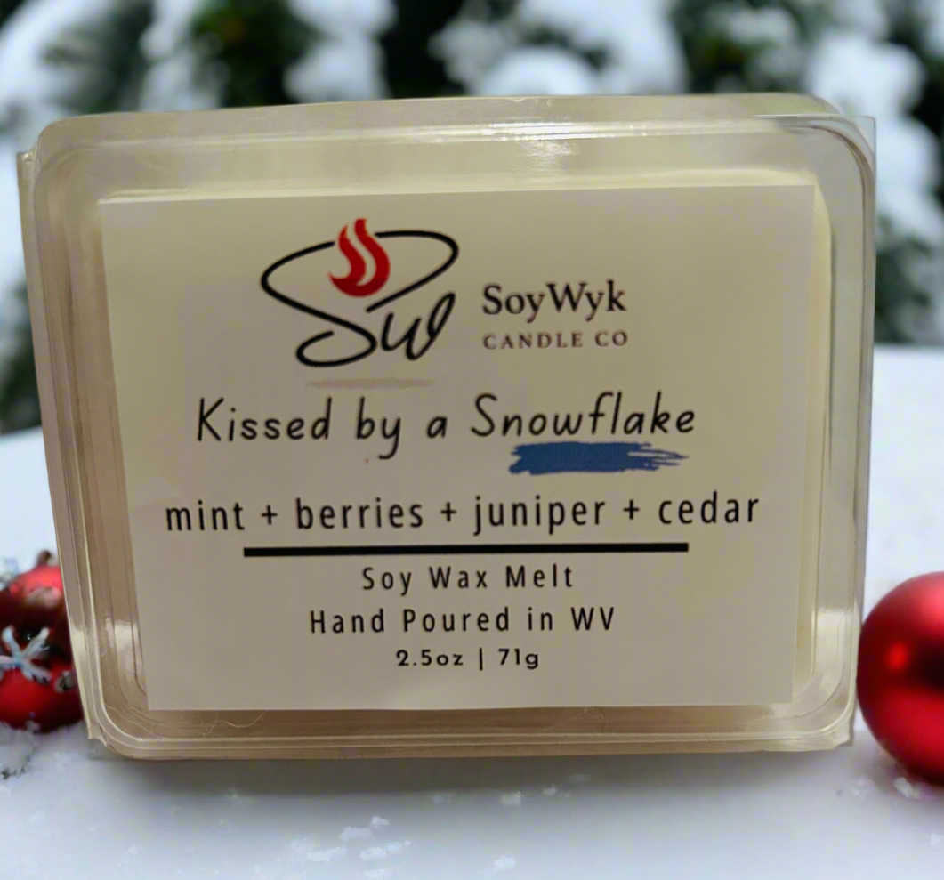 White Soy wax melt in clear 2.5 oz package.  Labeled “Kissed by a Snowflake “. Featured fragrant notes are mint, berries, juniper, cedar. In a white surface with red Christmas tree balks and green pine trees in background. Hand- poured in WV by SoyWyk Candle Co. 