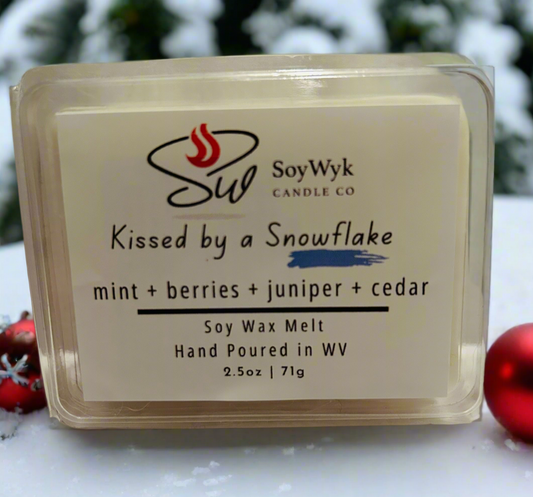 White Soy wax melt in clear 2.5 oz package.  Labeled “Kissed by a Snowflake “. Featured fragrant notes are mint, berries, juniper, cedar. In a white surface with red Christmas tree balks and green pine trees in background. Hand- poured in WV by SoyWyk Candle Co. 