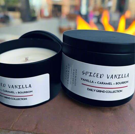 A candle in a black tin labeled “Spiced Vanilla “ is shown with featured fragrance notes of Vanilla Caramel and Bourbon. The background is a faded science of a living room with flames from fireplace showing behind the candles. Hand- poured in WV by SoyWyk Candle Co. 