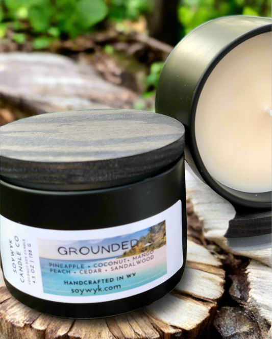 White Coconut Soy wax candle in modern black 6 oz tin  with wood lid labeled "Grounded" in an outside setting on a wood surface surrounded by greenery. Featured fragrance noted are pineapple, coconut, mango, peach, cedar, sandalwood.This is one of the Signature Collection from Soywyk Candle Co. hand-poured in WV.