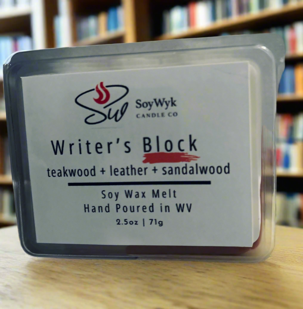White Soy wax melt in 2.5 ox clear package labeled “ Writers Block” Featured fragrant notes are  teakwood, leather, sandalwood sitting on a wood desk in a library surrounded by books on shelves. Hand-poured in WV by SoyWyk Candle Co. 