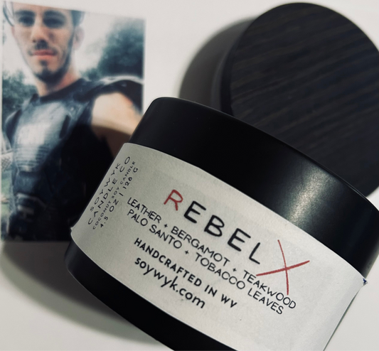 White Coconut Soy Candle in a modern black reusable ceramic jar with a wood lid labeled "Rebel" Featured fragrance notes are leather, bergamot, teakwood, palo Santo, tobacco leaves on a white background with a young guy in motorcycle gear in the background. One of the Signature Collections from Soywyk Candle Co hand-poured in WV.