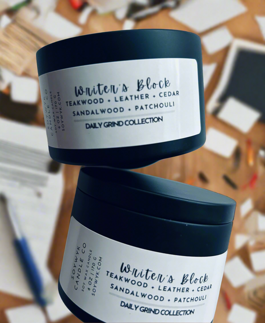 A candle in a black modern tin labeled “Writers Block” is shown from the SoyWyk Daily Grind Collection. Featured fragrant notes are teakwood, leather, sandalwood. On a faded background with books, papers and pens scattered randomly about. Hand poured in WV by SoyWyk  Candle Co. 