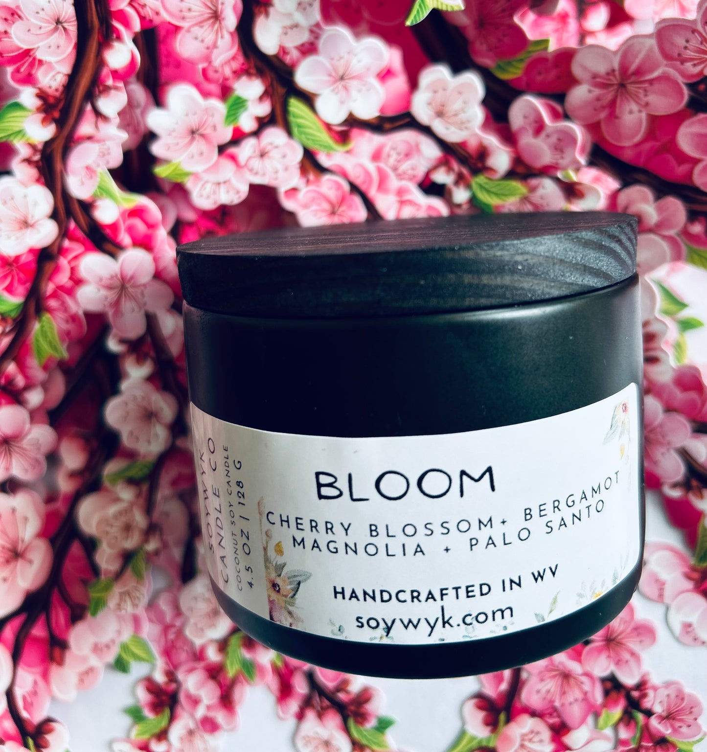 Coconut Soy Candle in a 5oz modern black reusable ceramic jar with a wood lid labeled "Bloom". Sitting in the middle a cherry blossom background .Featured fragrance notes are cherry blossoms, bergamot,manola, palo Santo. One  of the Signature Collections from Soywyk Candle Co.  hand-poured in WV.