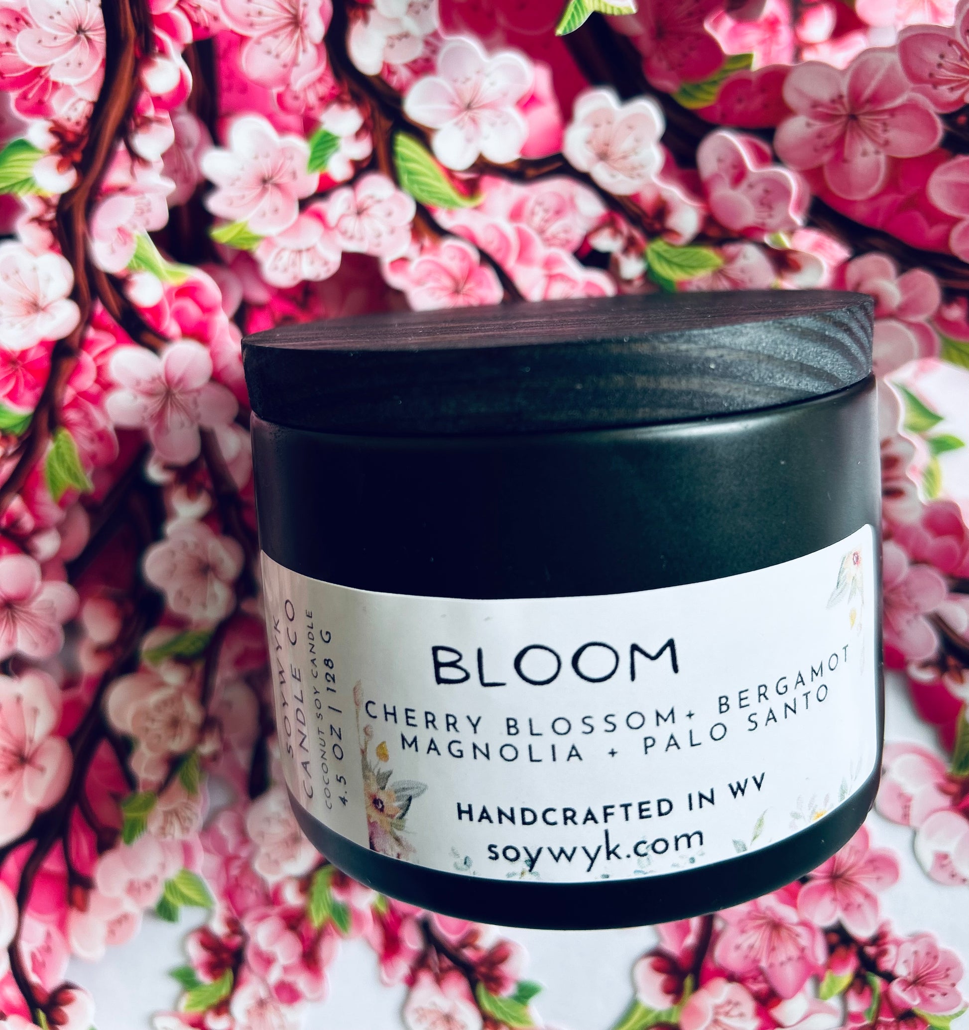 Coconut Soy Candle in a 5oz modern black reusable ceramic jar with a wood lid labeled "Bloom". Sitting in the middle a cherry blossom background .Featured fragrance notes are cherry blossoms, bergamot,manola, palo Santo. One  of the Signature Collections from Soywyk Candle Co.  hand-poured in WV.