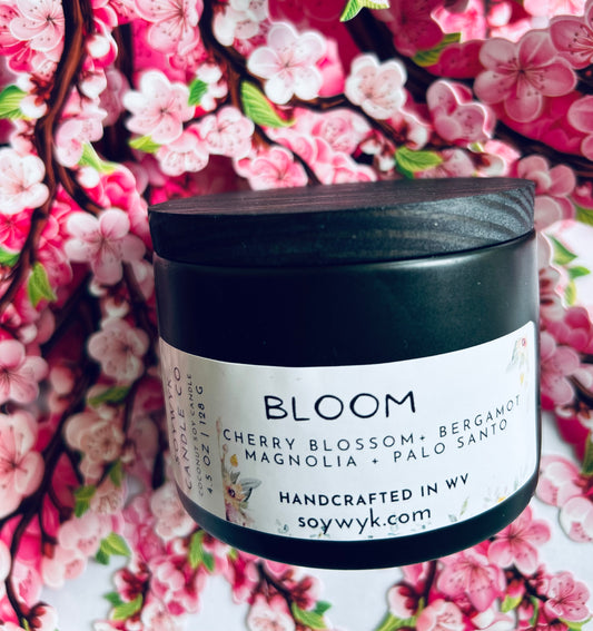 Coconut Soy Candle in a 5oz modern black reusable ceramic jar with a wood lid labeled "Bloom". Sitting in the middle a cherry blossom background .Featured fragrance notes are cherry blossoms, bergamot,manola, palo Santo. One  of the Signature Collections from Soywyk Candle Co.  hand-poured in WV.