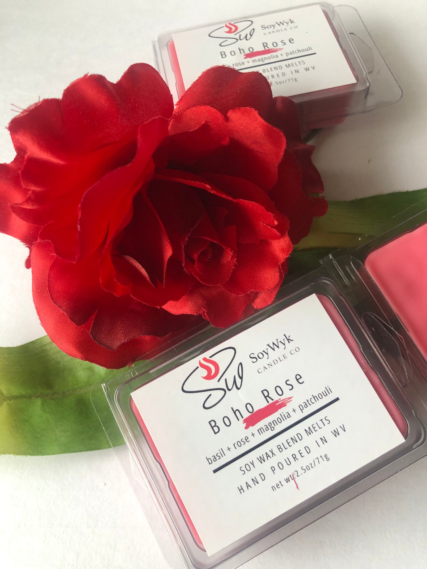 Red Soy wax melt in 2.5 oz clear package labeled “Boho Rose" on a white counter isurrounded by a rose and  green leaves. Featured fragrant notes are  basil, rose, magnolia, patchouli. Hand- poured in WV by SoyWyk Candle Co. 