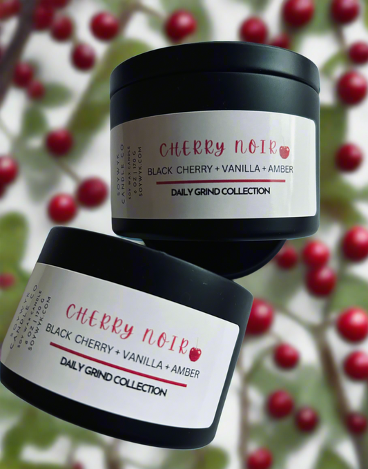 Soy candle in a modern black 6 oz candle tin labeled "Cherry Noir" on background surrounded by red cherries and green leaves. Featured fragrance notes are black cherry, vanilla, amber. One of the Daily Grind Collection by Soywyk Candle Co. hand-poured in WV.
