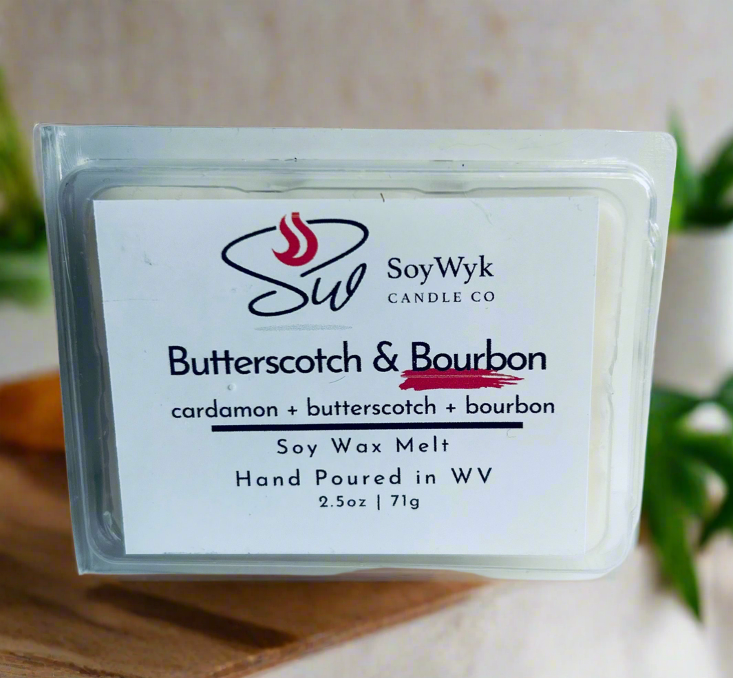 White Soy wax melt in 2.5 oz clear package labeled “Butterscotch and Bourbon on a wood counter 
in kitchen surrounded by green plants. Featured fragrant notes are  cardamon, butterscotch, bourbon. . Hand- poured in WV by SoyWyk Candle Co.