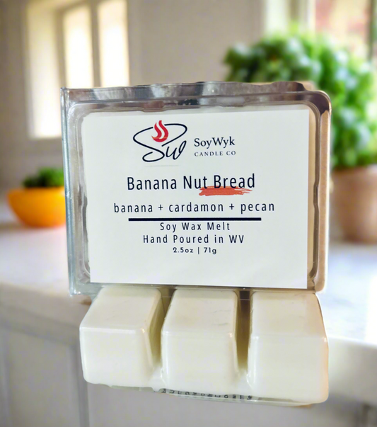 White Soy wax melt in 2.5 oz clear package labeled “Banana Nut Bread"  on a white ceramic countertop in kitchen  surrounded by green plants. Featured fragrant notes are  banana, cardamon, pecan. Hand- poured in WV by SoyWyk Candle Co. 