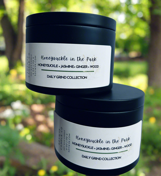 Soy wax candle in a modern black 6 oz tin labeled "Honeysuckle in the Park". In a park like setting surrounded by trees and flowers. Featured fragrance notes are  honeysuckle, jasmine, ginger, wood. One of the Daily Grind Collection fromSoywyk Candle Co. Hand-poured in WV. 