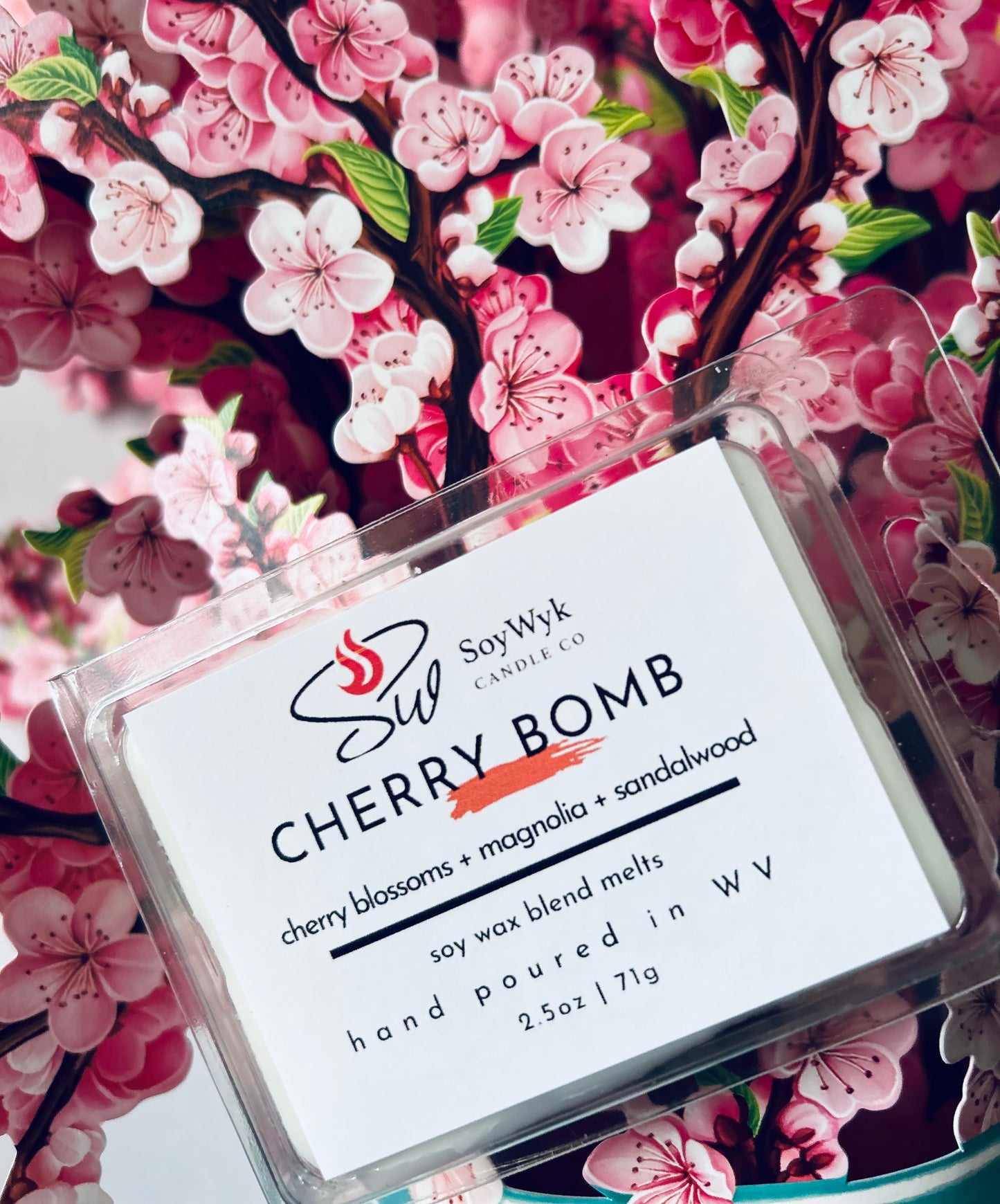 White Soy Wax Melt in a 2.5 oz clear package in a kitchen on a modern kitchen by a window with plants. Labeled as “Cherry Bomb” Featured fragrant notes are cherry blossoms, magnolia, sandalwood. Positioned in the middle of a bunch if pink cherry blossoms. 
Hand-poured in WV by SoyWyk Candle Co. 