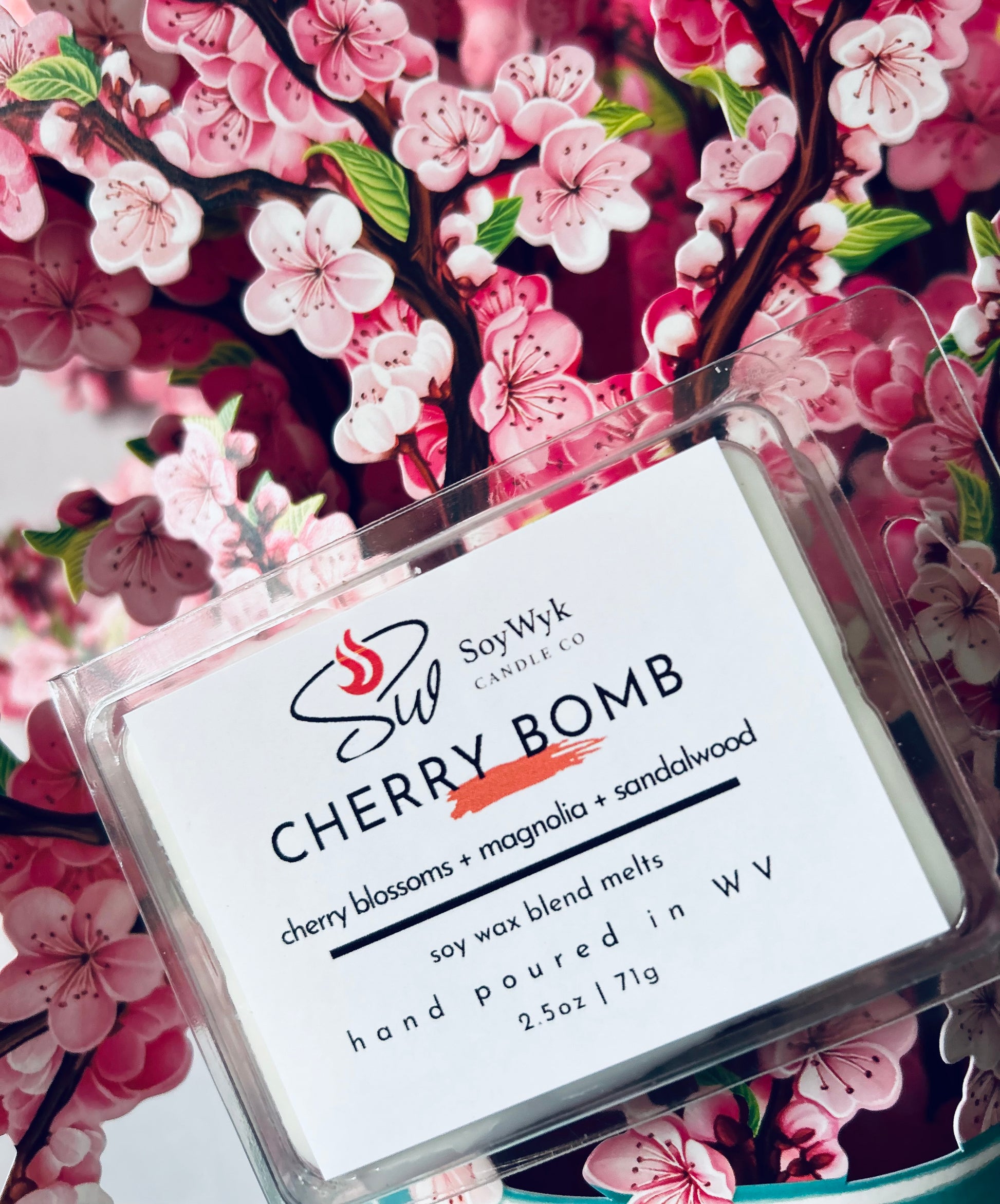 White Soy Wax Melt in a 2.5 oz clear package in a kitchen on a modern kitchen by a window with plants. Labeled as “Cherry Bomb” Featured fragrant notes are cherry blossoms, magnolia, sandalwood. Positioned in the middle of a bunch if pink cherry blossoms. 
Hand-poured in WV by SoyWyk Candle Co. 