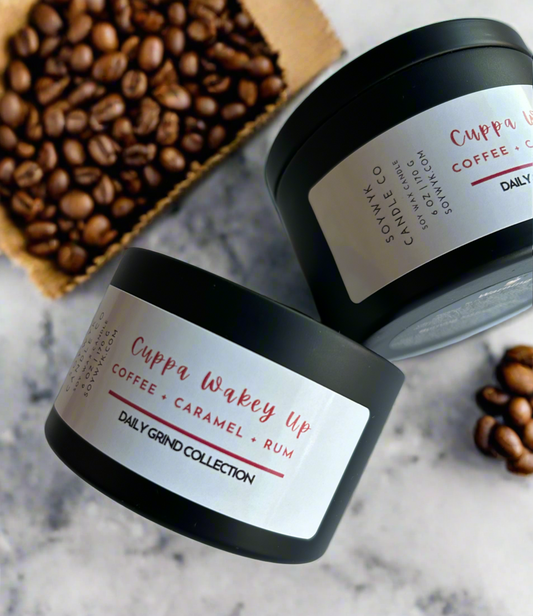 White Soy Candle in a 6 oz modern black tin labeled “Cuppa Wakey Up” Featured fragrant notes are coffee, caramel, rum on a white ceramic counter top in the kitchen with a scoop of coffee  beans beside it.  One of the Daily Grind Collections   Hand-poured from SoyWyk Candle Co. 
